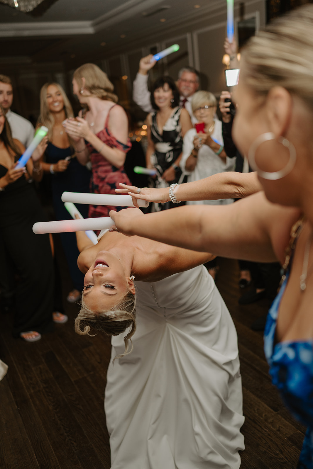 Bride laughs while doing limbo under glow sticks held by guests on the lively reception dance floor.